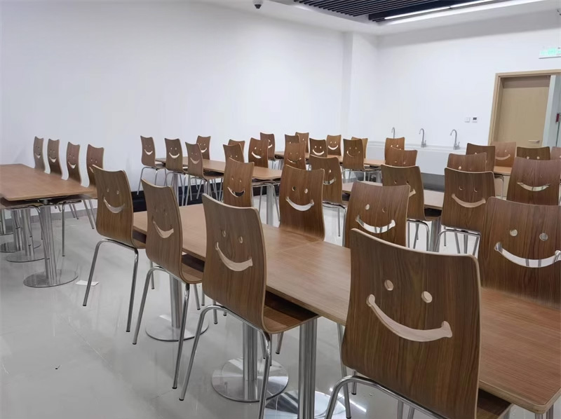 Smile Backrest Plywood Canteen Chair2 Spacious canteen furnished with rows of smile backrest wooden canteen chairs and rectangular dining tables, ideal for schools or company cafeterias