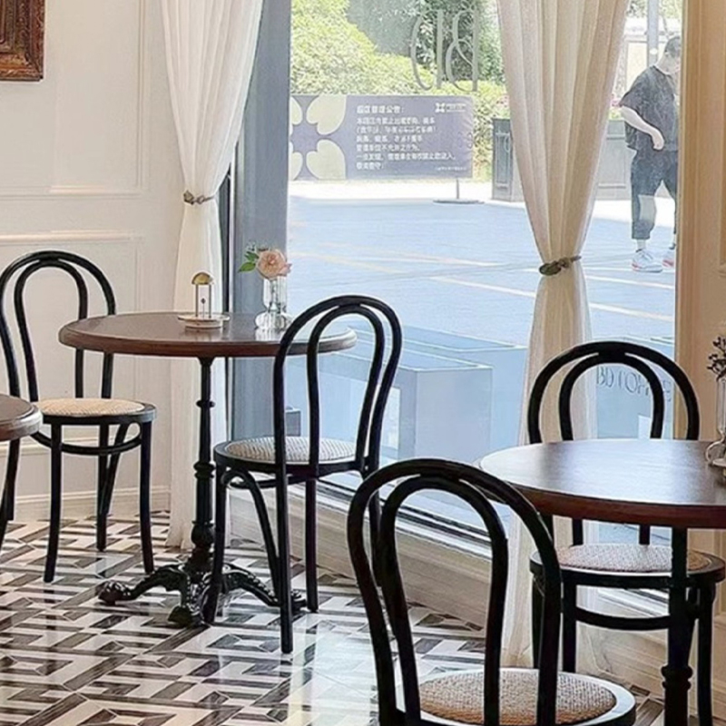 Multiple vintage café tables with wooden tops and black bentwood chairs near a window.