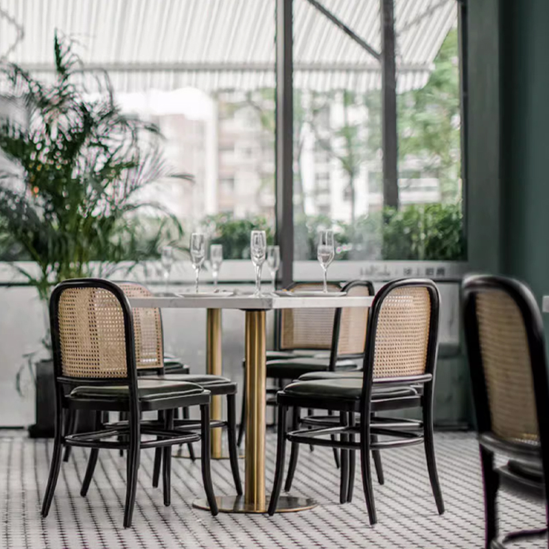 Vintage Rattan Back Restaurant Chair1 Contemporary dining area featuring black rattan back dining chairs with green leather seats and gold base tables, surrounded by large glass windows and indoor plants